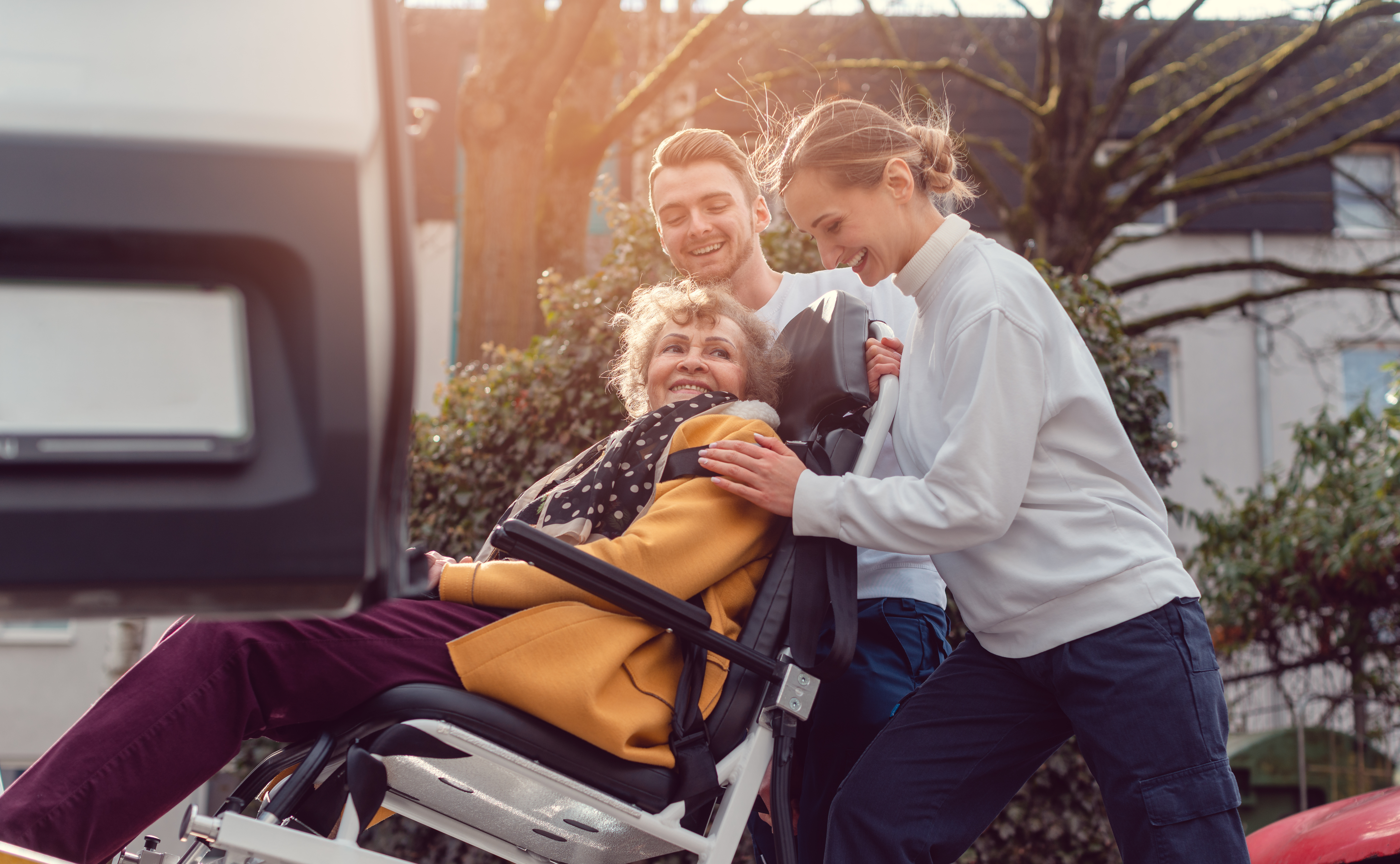 Smiling older woman in a wheelchair being assisted by two caregivers near a transport vehicle.