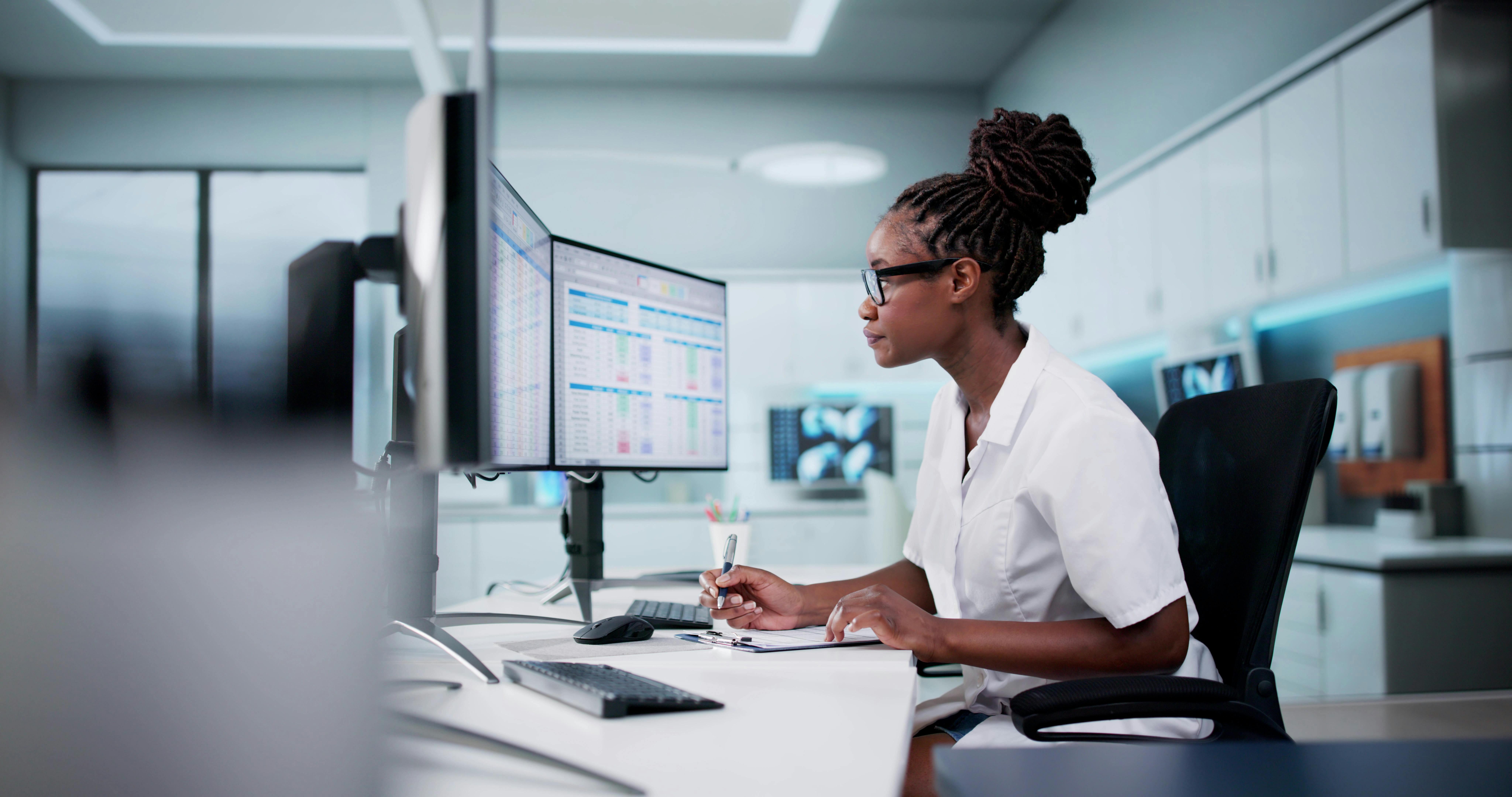 Healthcare professional working at dual computer monitors with spreadsheets in a modern office.