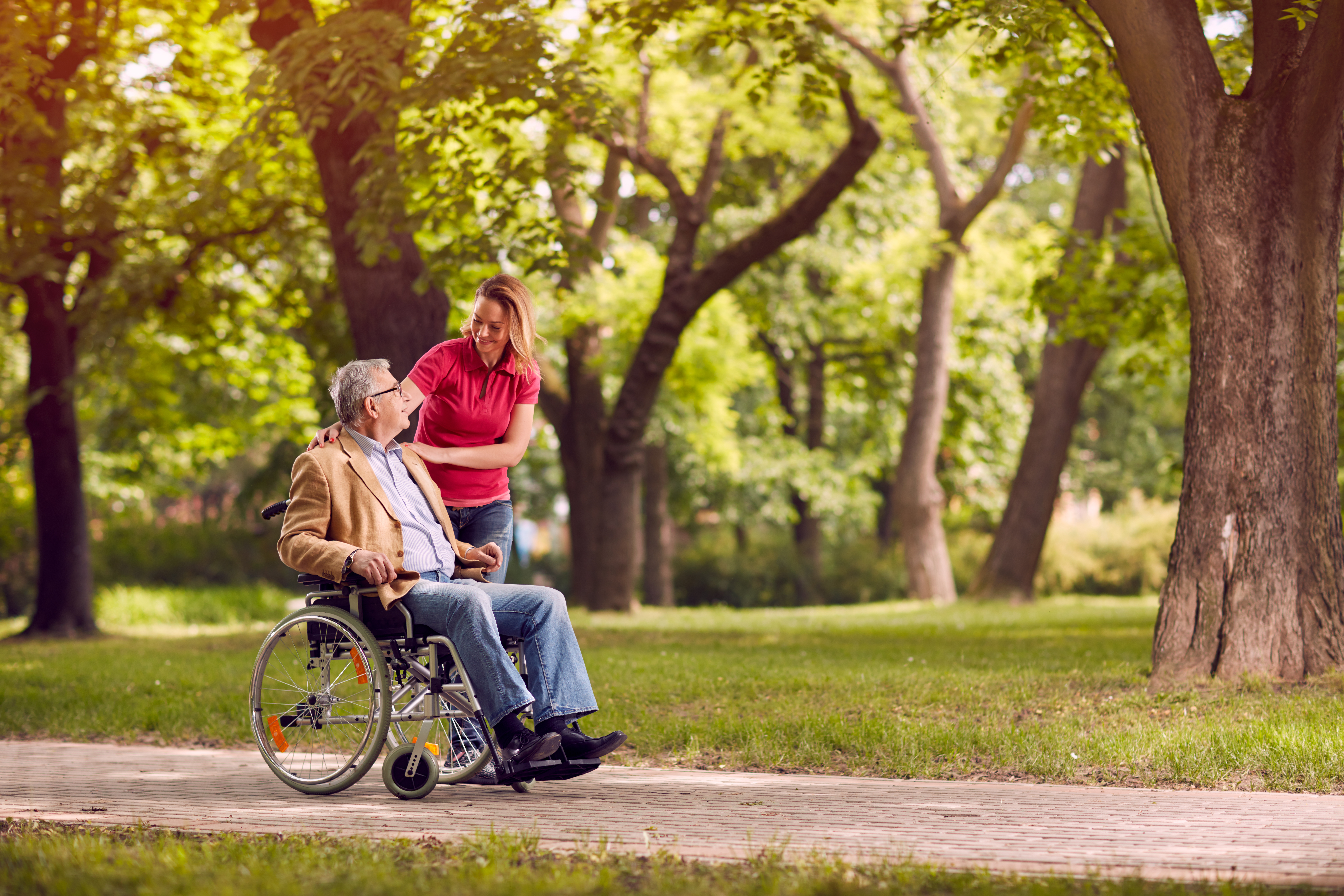Smiling woman assisting an older man in a wheelchair along a park pathway.