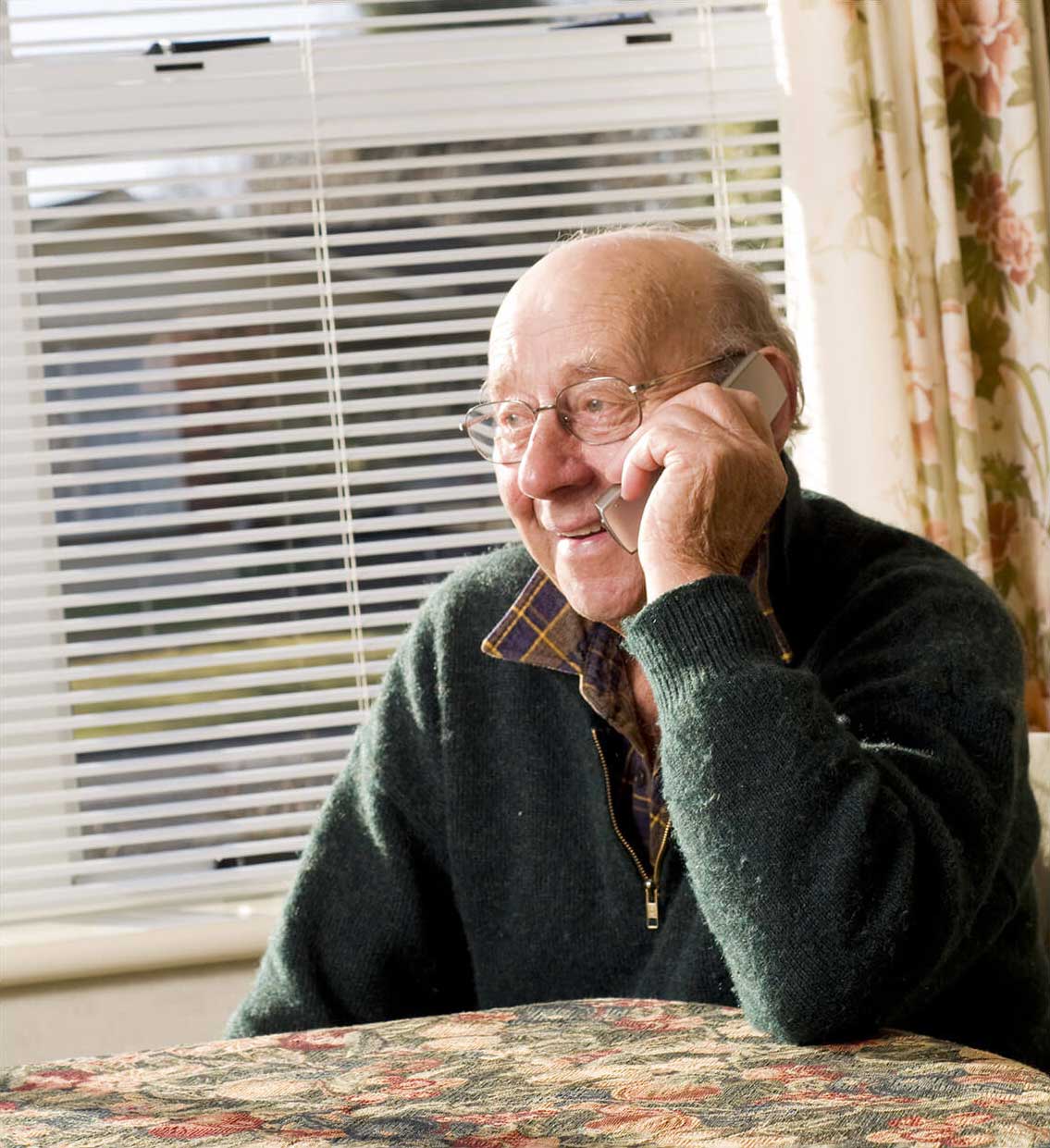 Smiling older man sitting at a table and talking on the phone at home.