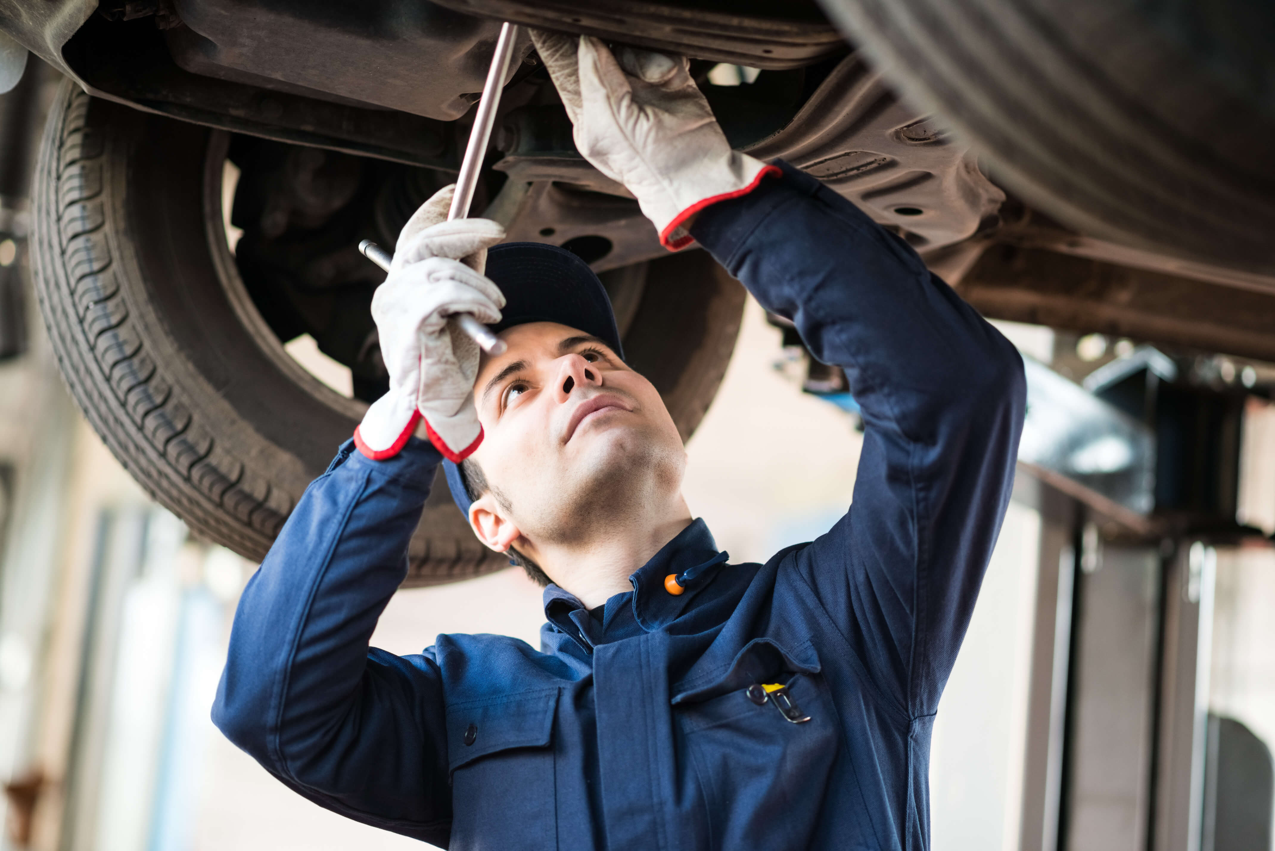 Auto mechanic in uniform using tools to work underneath a lifted vehicle.