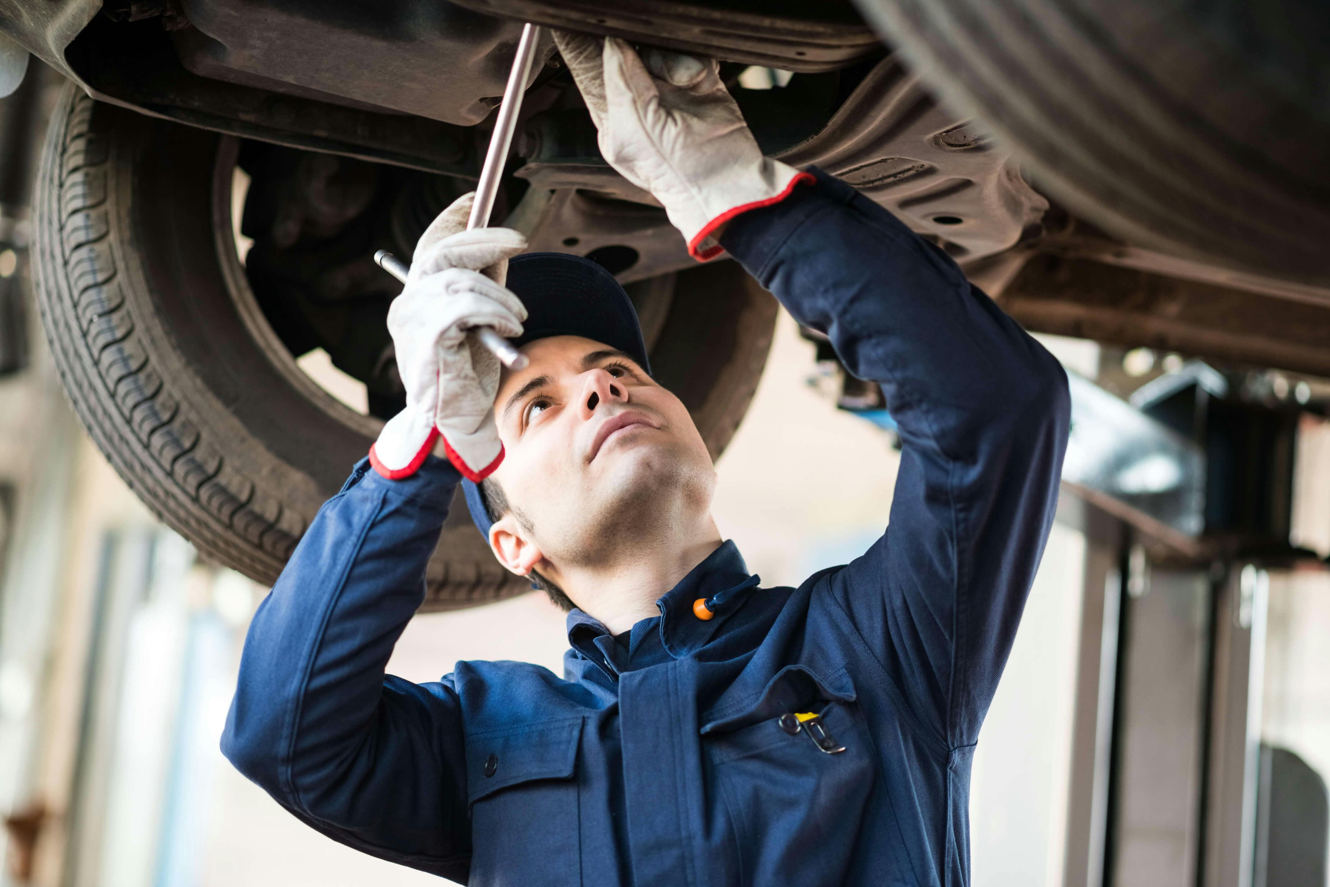 Auto mechanic in uniform using tools to work underneath a lifted vehicle.