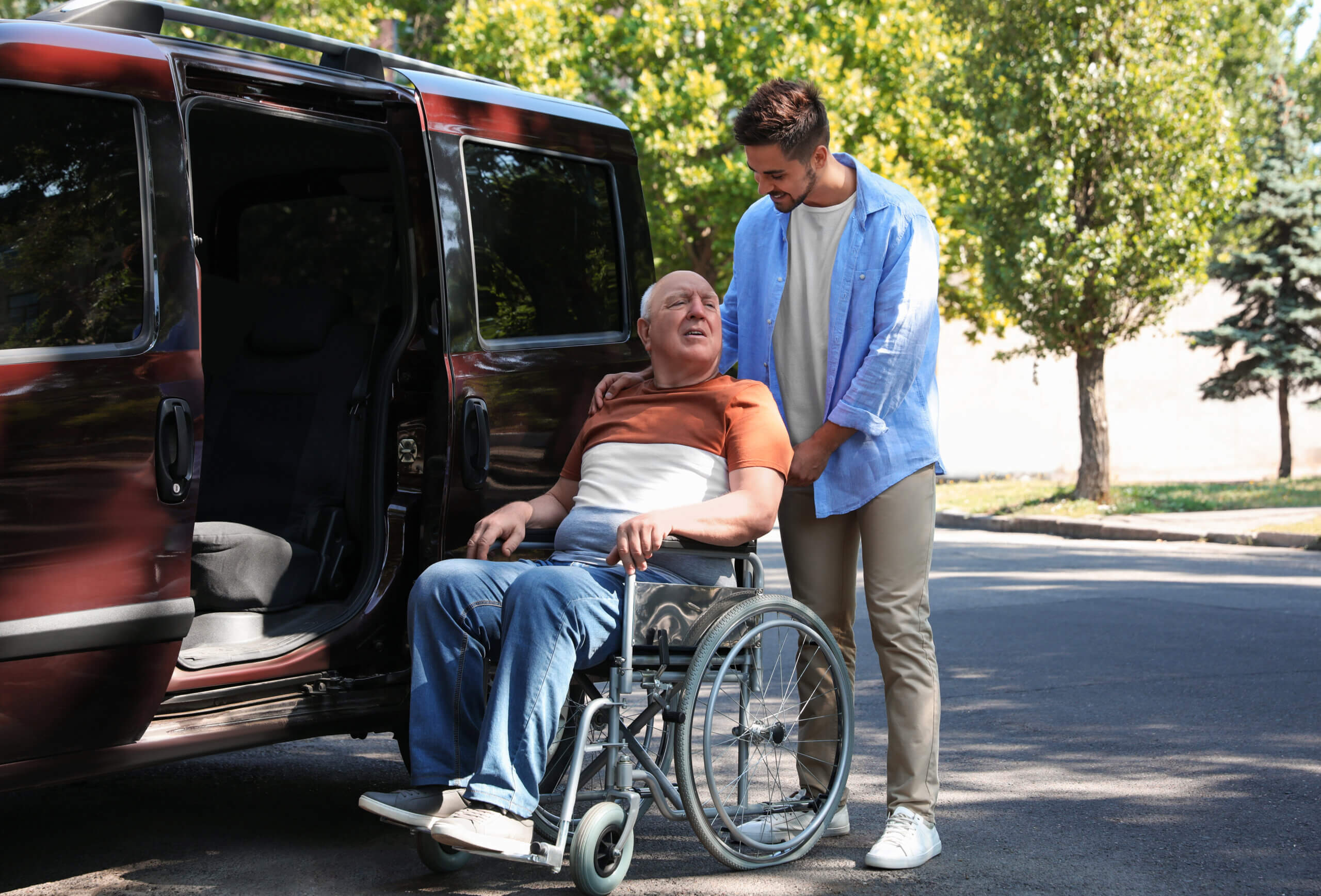 Smiling man assisting an older man in a wheelchair next to a transport van.