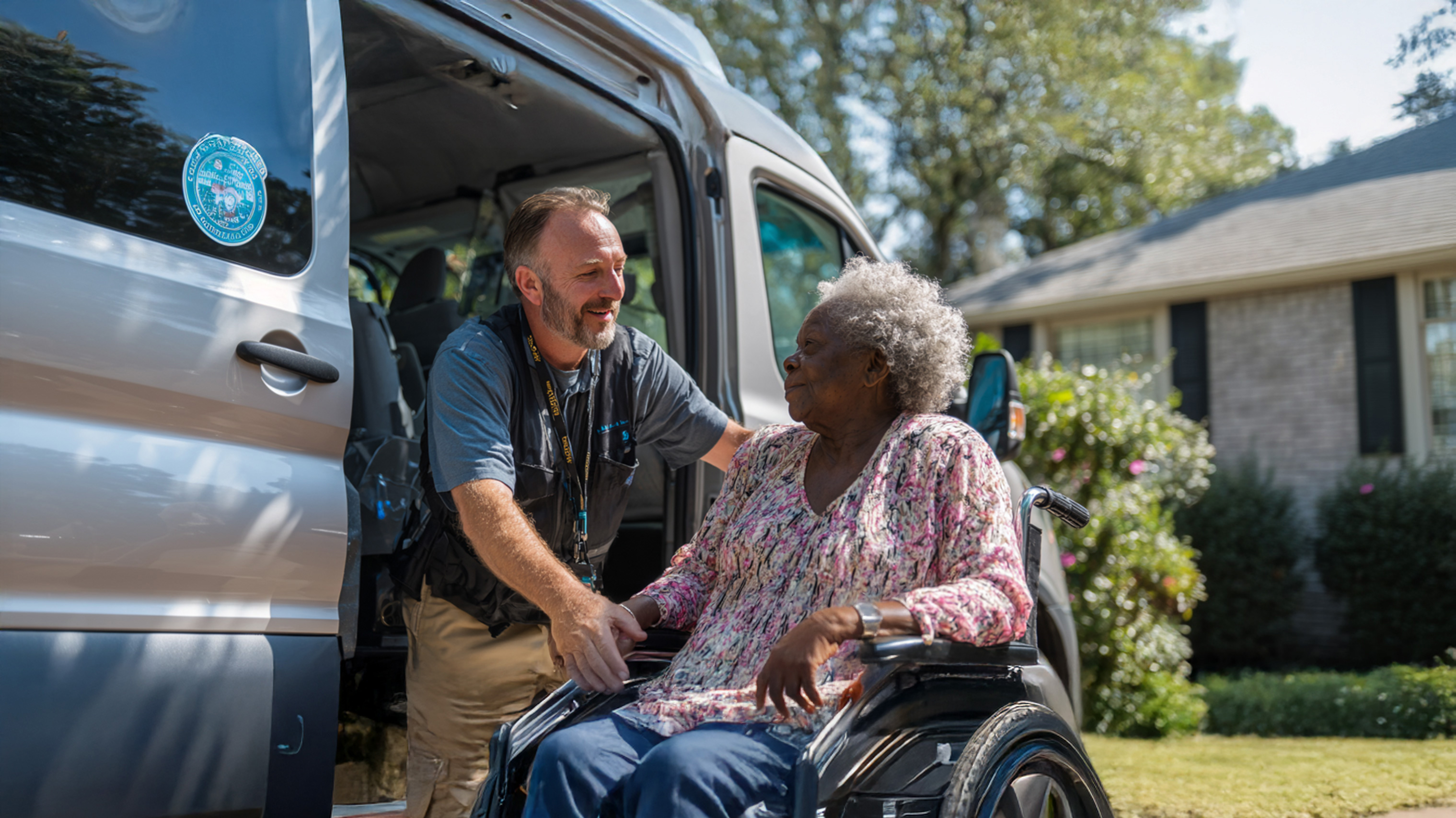 A transportation worker assists an elderly woman in a wheelchair as she exits a van parked in front of a suburban home on a sunny day.