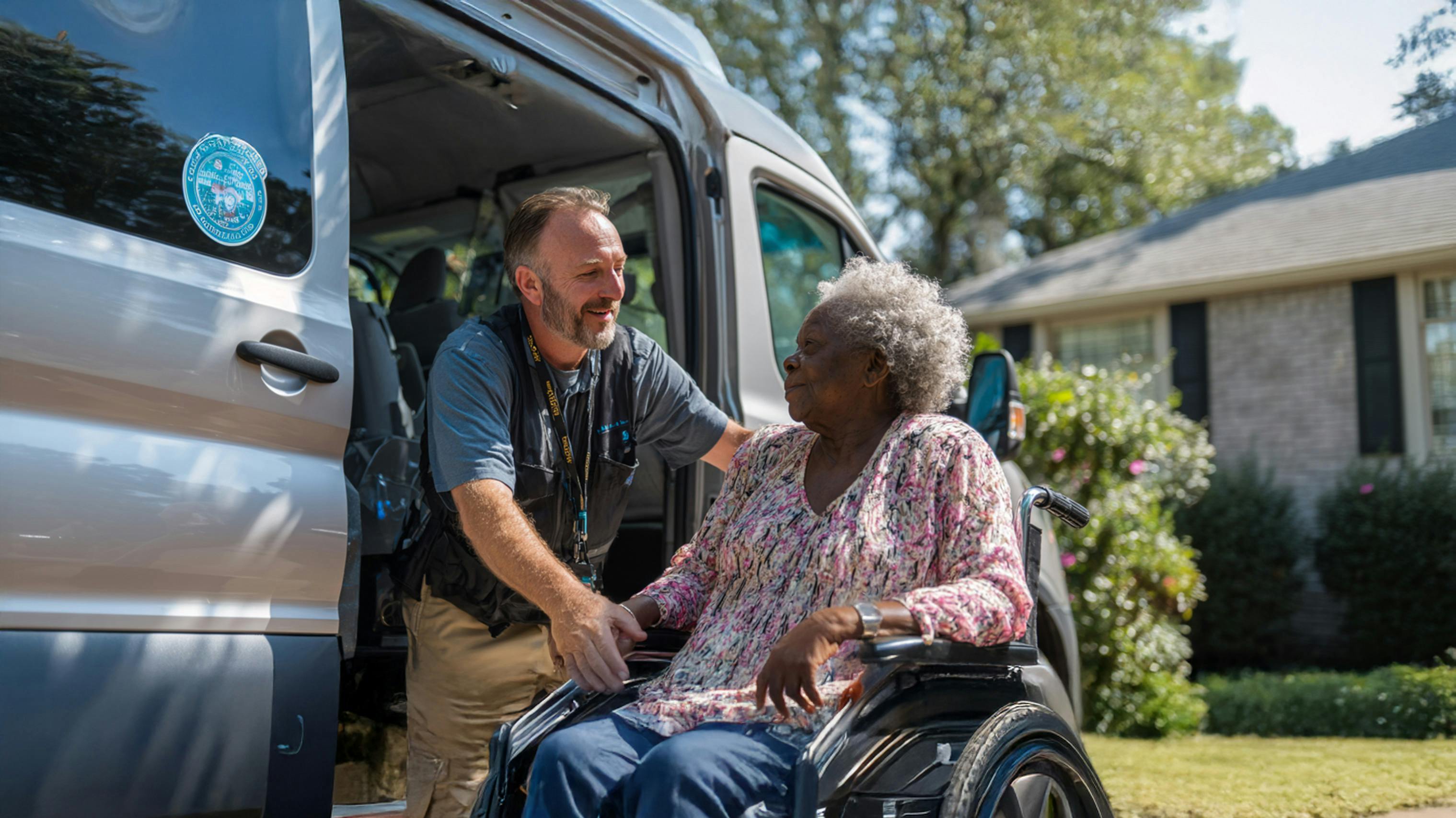 A transportation worker assists an elderly woman in a wheelchair as she exits a van parked in front of a suburban home on a sunny day.