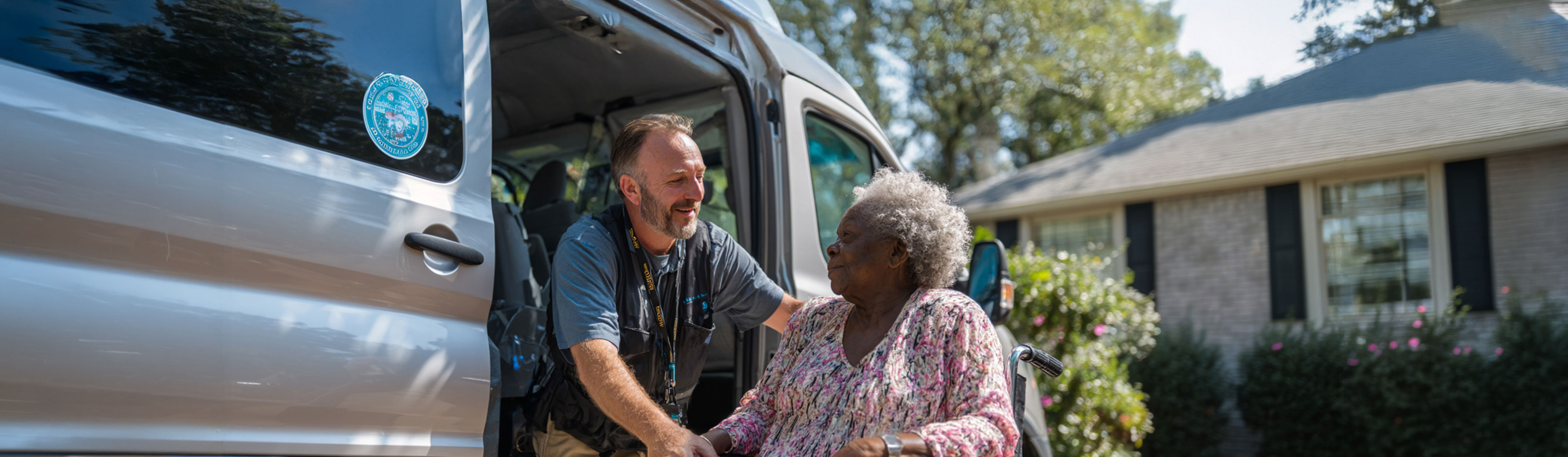 A transportation worker assists an elderly woman in a wheelchair as she exits a van parked in front of a suburban home on a sunny day.