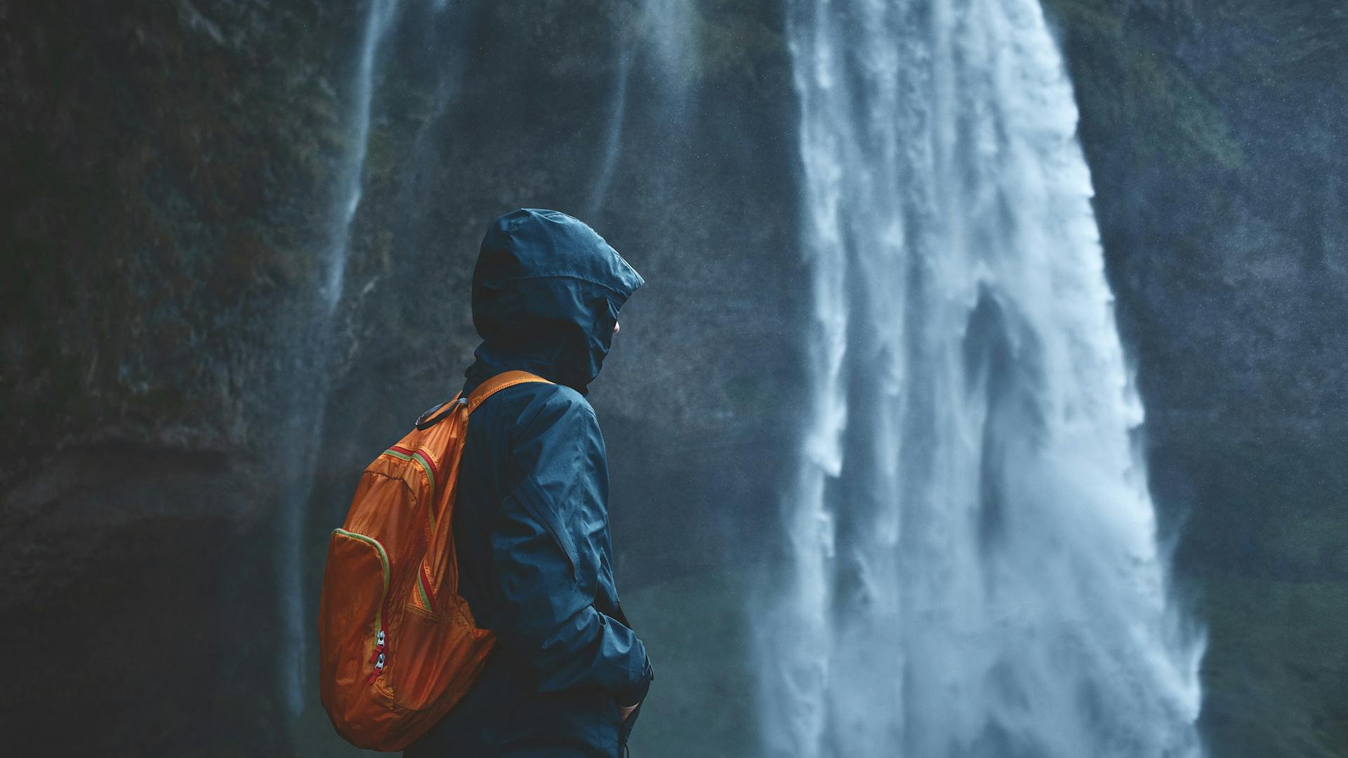 A person using a blue jacket and an orange backpack with a waterfall in the background.