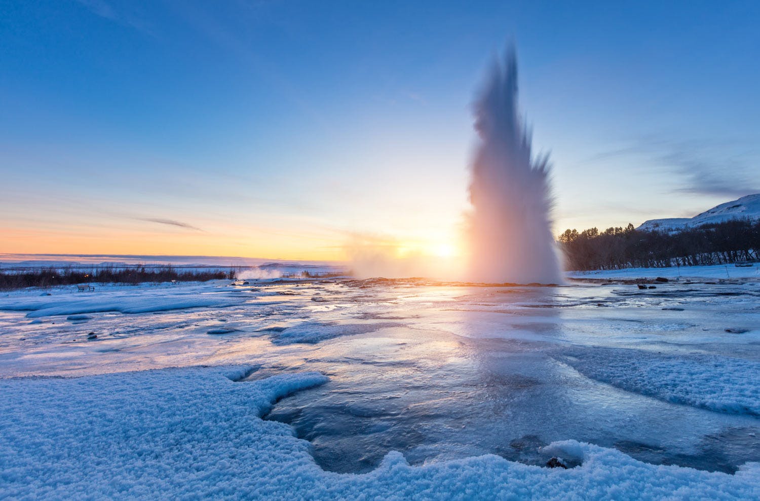 Geysir spewing boiling water into the air during sunset, all surroundings are frozen due to low temperatures in winter.