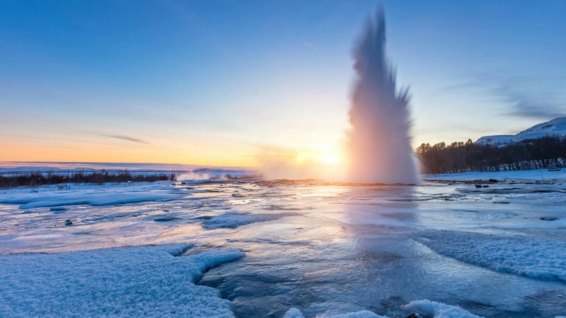 Geysir spewing boiling water into the air during sunset, all surroundings are frozen due to low temperatures in winter.