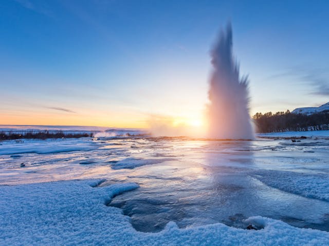 Geysir spewing boiling water into the air during sunset, all surroundings are frozen due to low temperatures in winter.