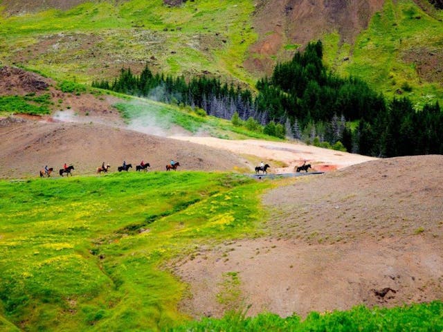 People are riding horses, and hot steam is coming up from the ground between green scenery and scorched earth.