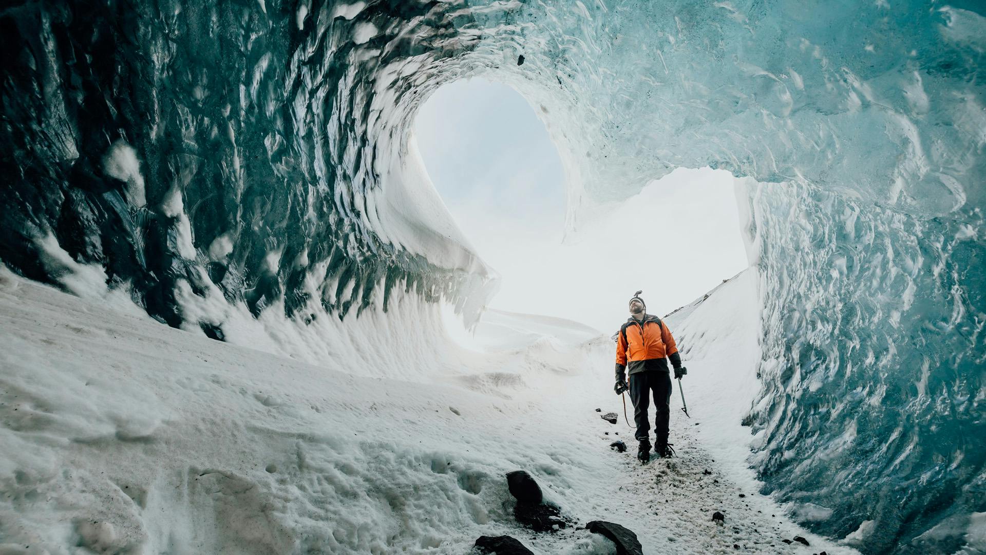 A person using an orange jacket is standing and looking at the ceiling of an ice cave.