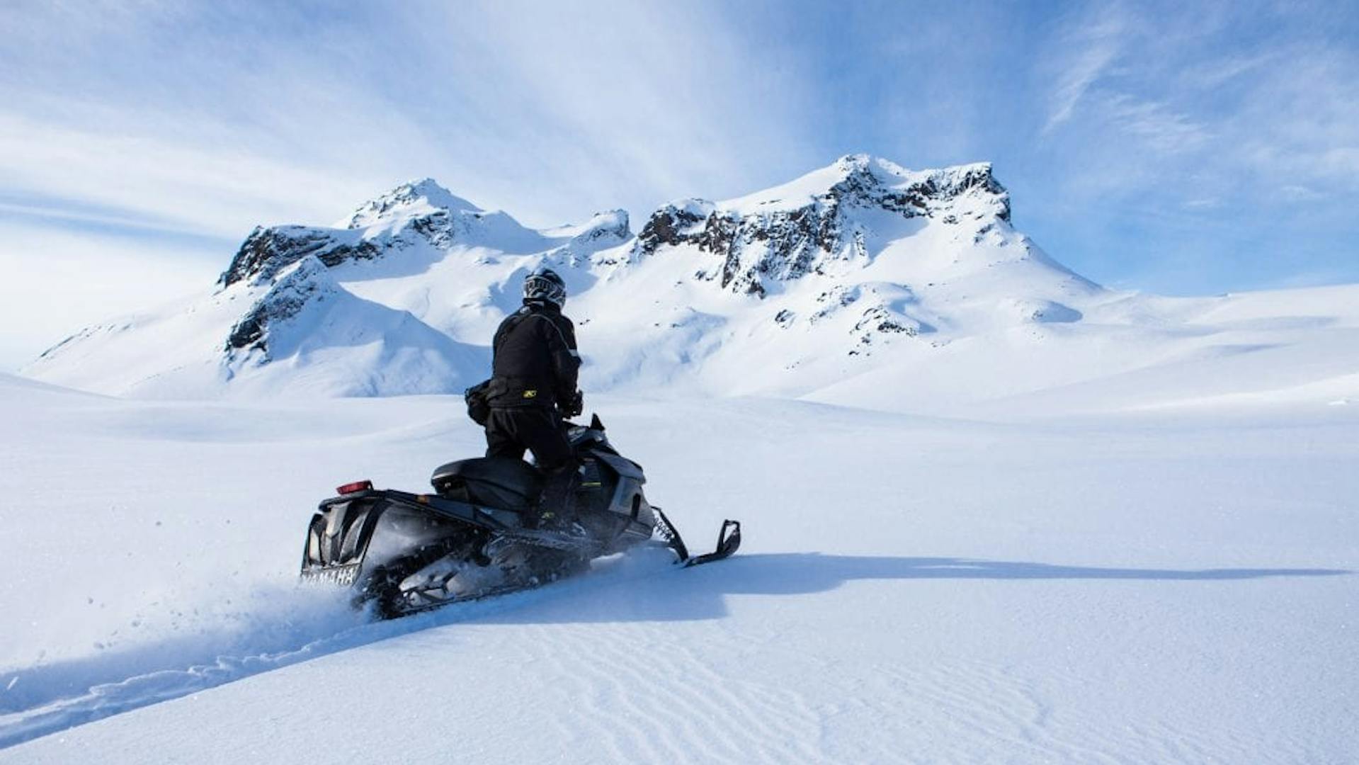 A man is standing while driving a snowmobile fast over an icy glacier toward a snowy mountaintop.