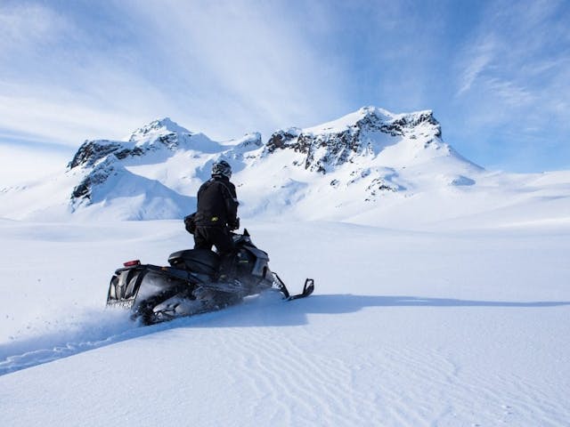 A man is standing while driving a snowmobile fast over an icy glacier toward a snowy mountaintop.