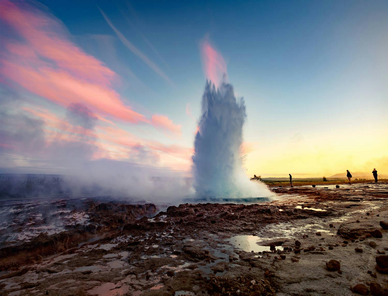 Geysir spewing boiling water in the air while people are standing in the surrounding to appreciate this natural phenomenom.