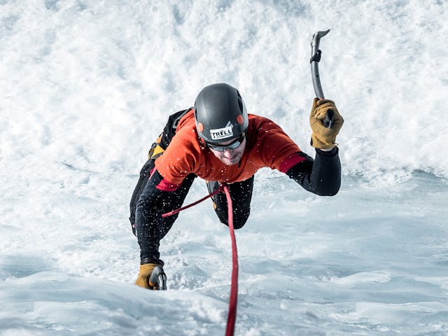 Man climbing an ice wall with two ice axes, the ice wall is covered with snow.