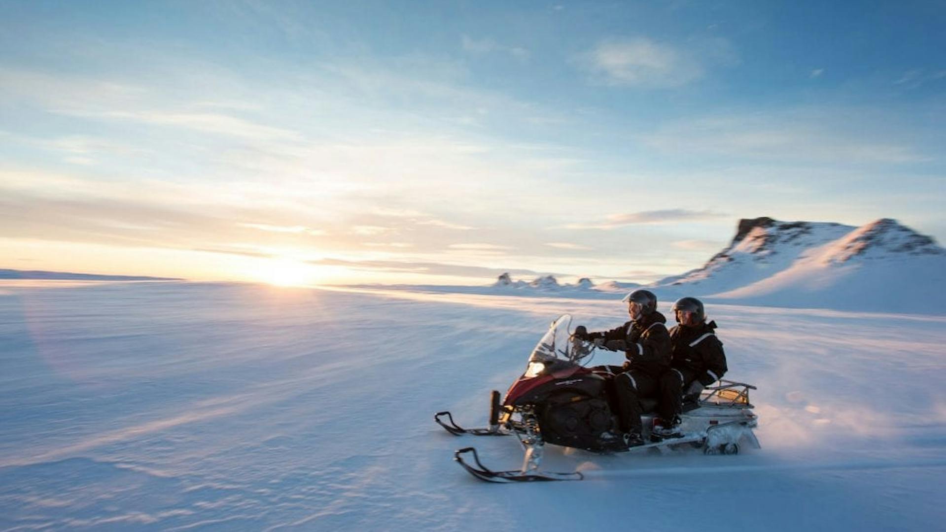 Two people share a snowmobile while they ride over a glacier; the sky is clear blue, the sun is coming up on the horizon, and mountain peaks are in the background.