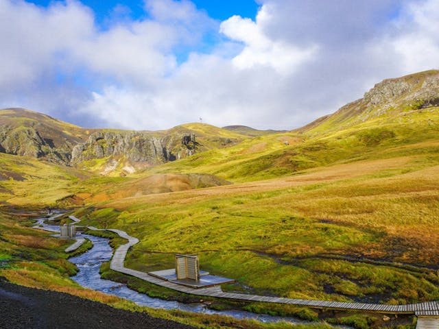 A warm geothermal river during summertime flows through a green valley in Reykjadalur, with a pathway for people to change clothes more efficiently.