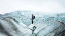 A man standing on top of an ice chunk, behind him it is possible to see glacier formations.