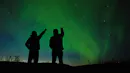 Silhouette of two person point to the sky, while an aurora borealis is showing up.