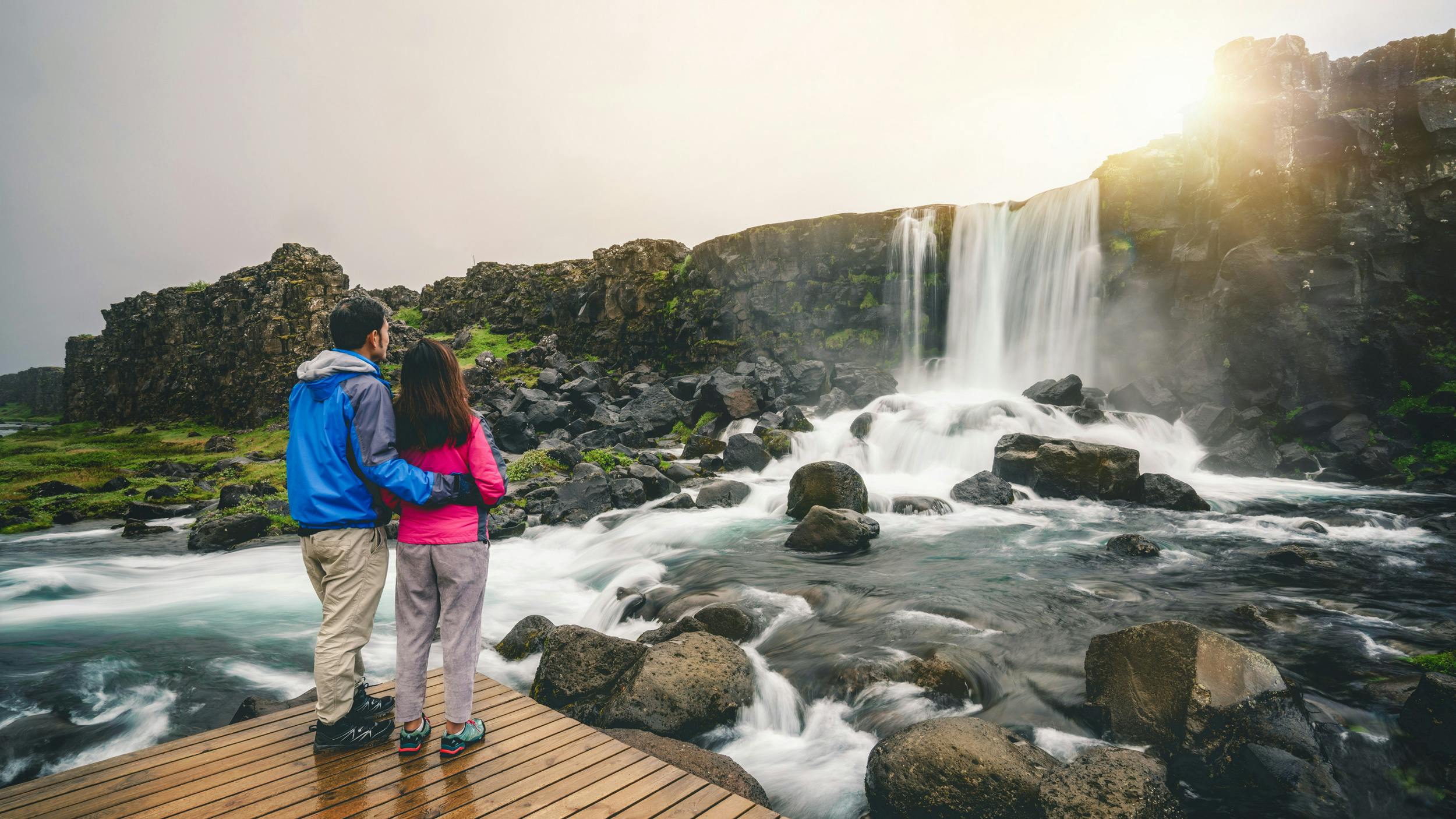 A couple holding each other while they are looking at a waterfall, situated in Thingvellir National Park.