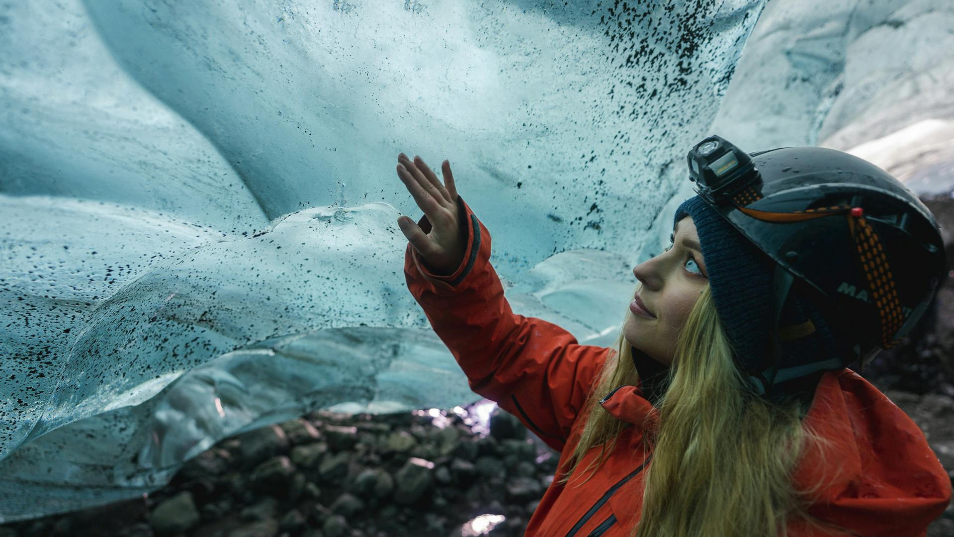 Blonde woman wearing a helmet with a flashlight touching the ice of an ice cave.