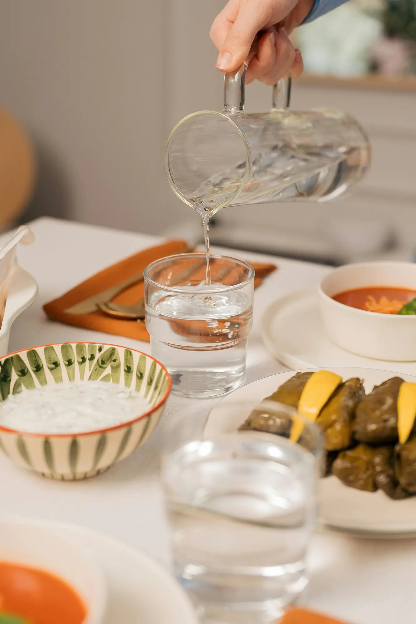 Water is being poured from a carafe into glasses on a kitchen table - the table is set with bowls, napkins, and silverware
