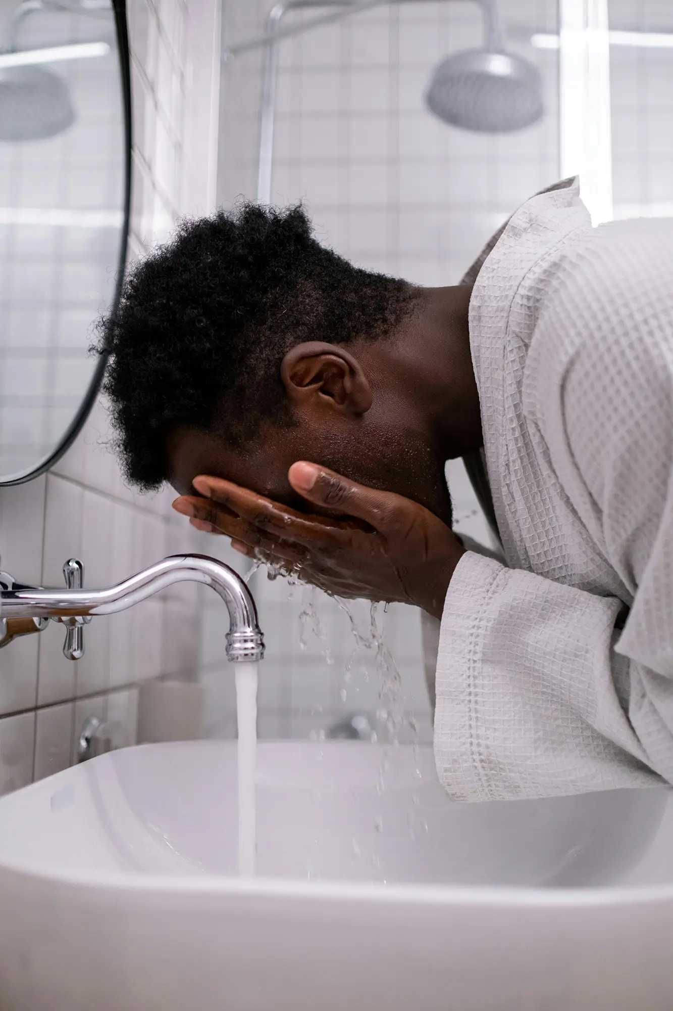 Young black person in a waffle-knit robe washing their face at a bathroom sink