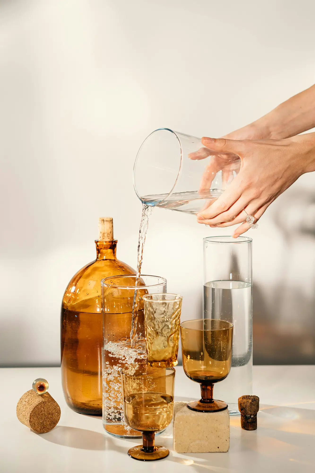 A pair of hands is pouring crystal clear water from a glass carafe into an artistically-arranged collection of clean and amber-colored vessels