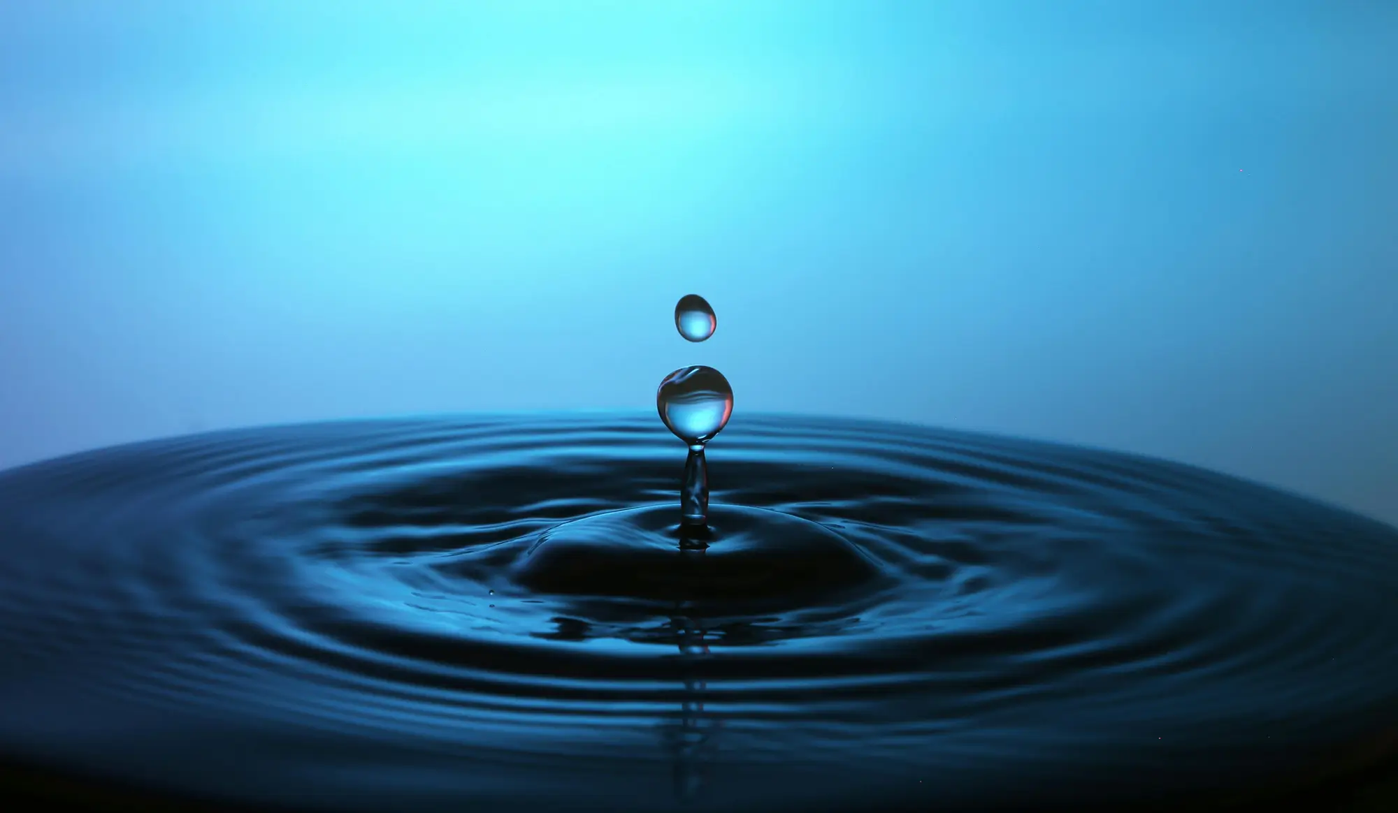Extreme closeup of a blue water droplet falling into a larger pool of water, causing it to ripple outwards