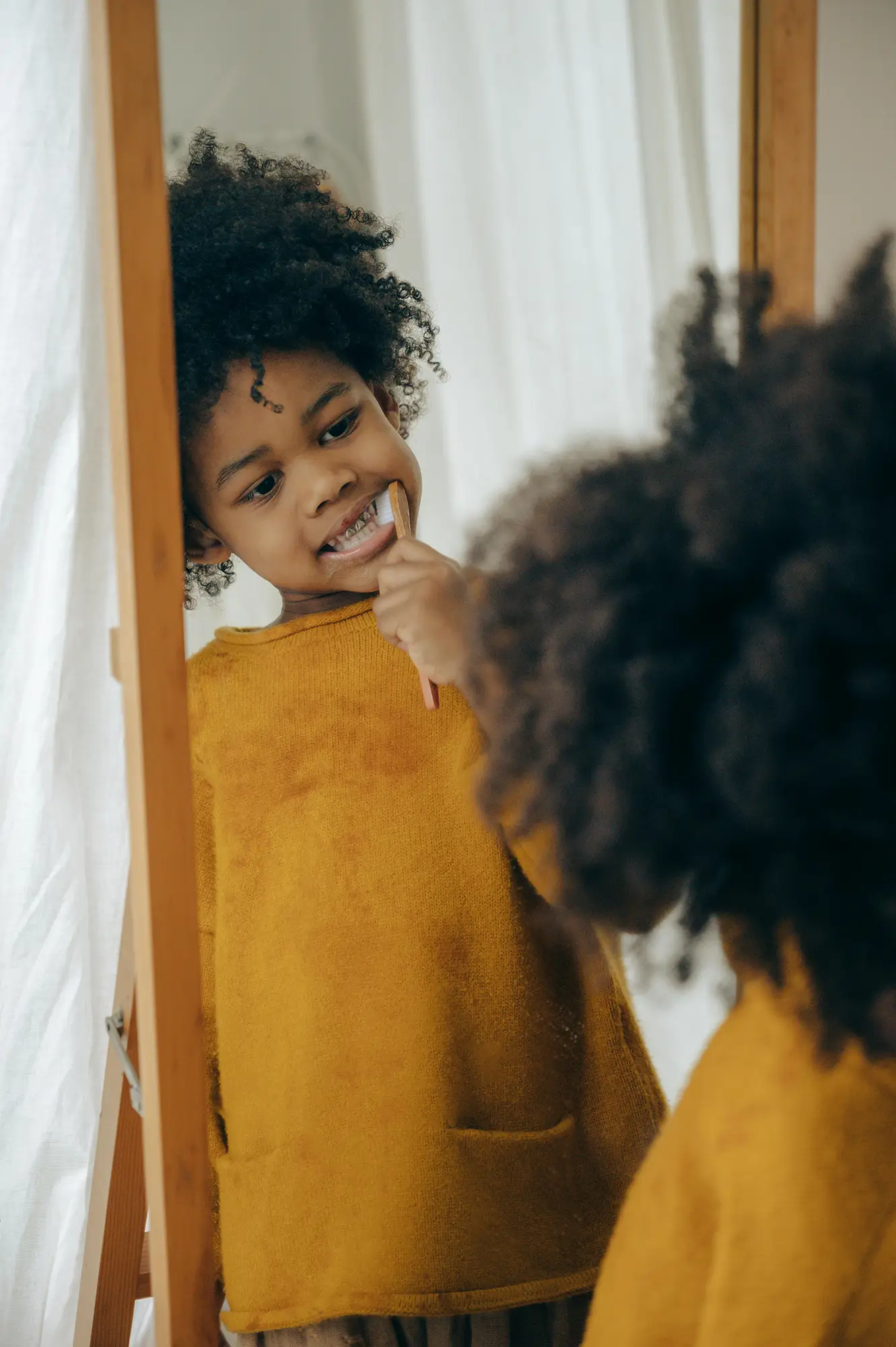 Young black child in a mustard yellow top brushing their teeth in front of a mirror