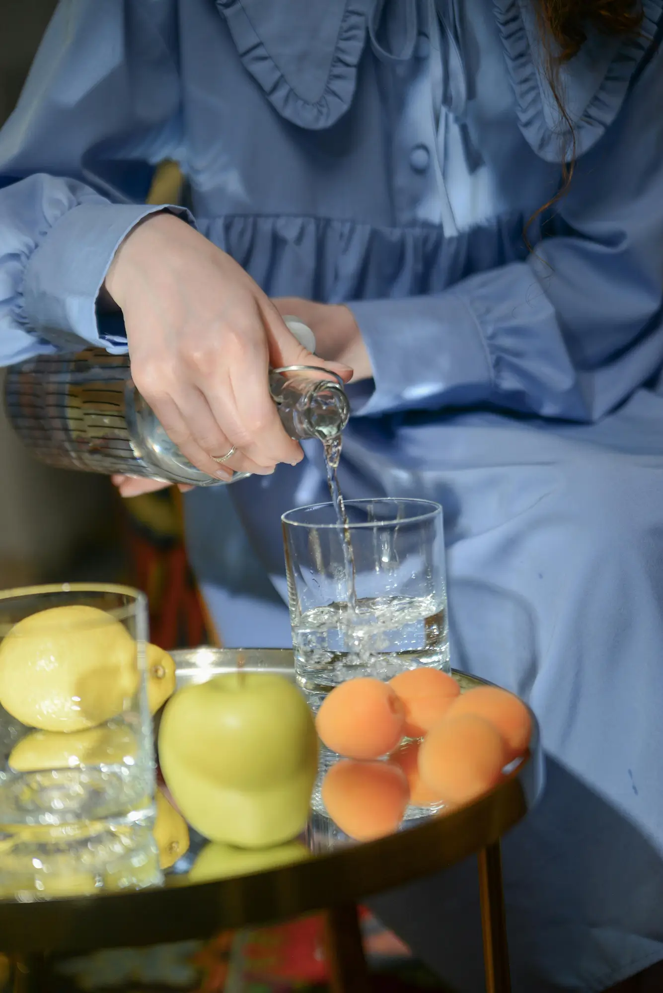 Person in a blue dress pouring water into a glass from a carafe - there is fresh fruit in the foreground