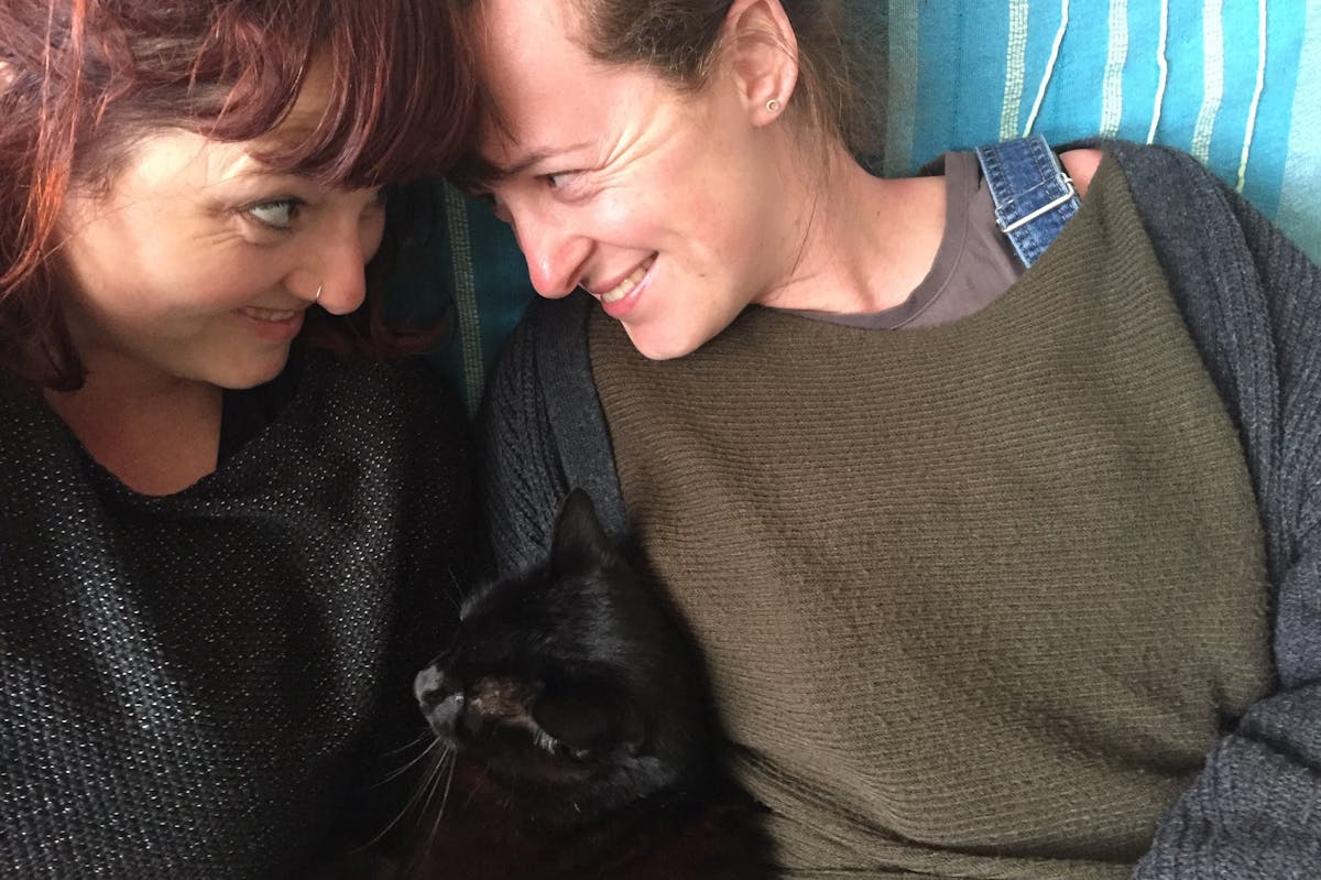 Two women looking at each other, with a black cat cuddled in-between them