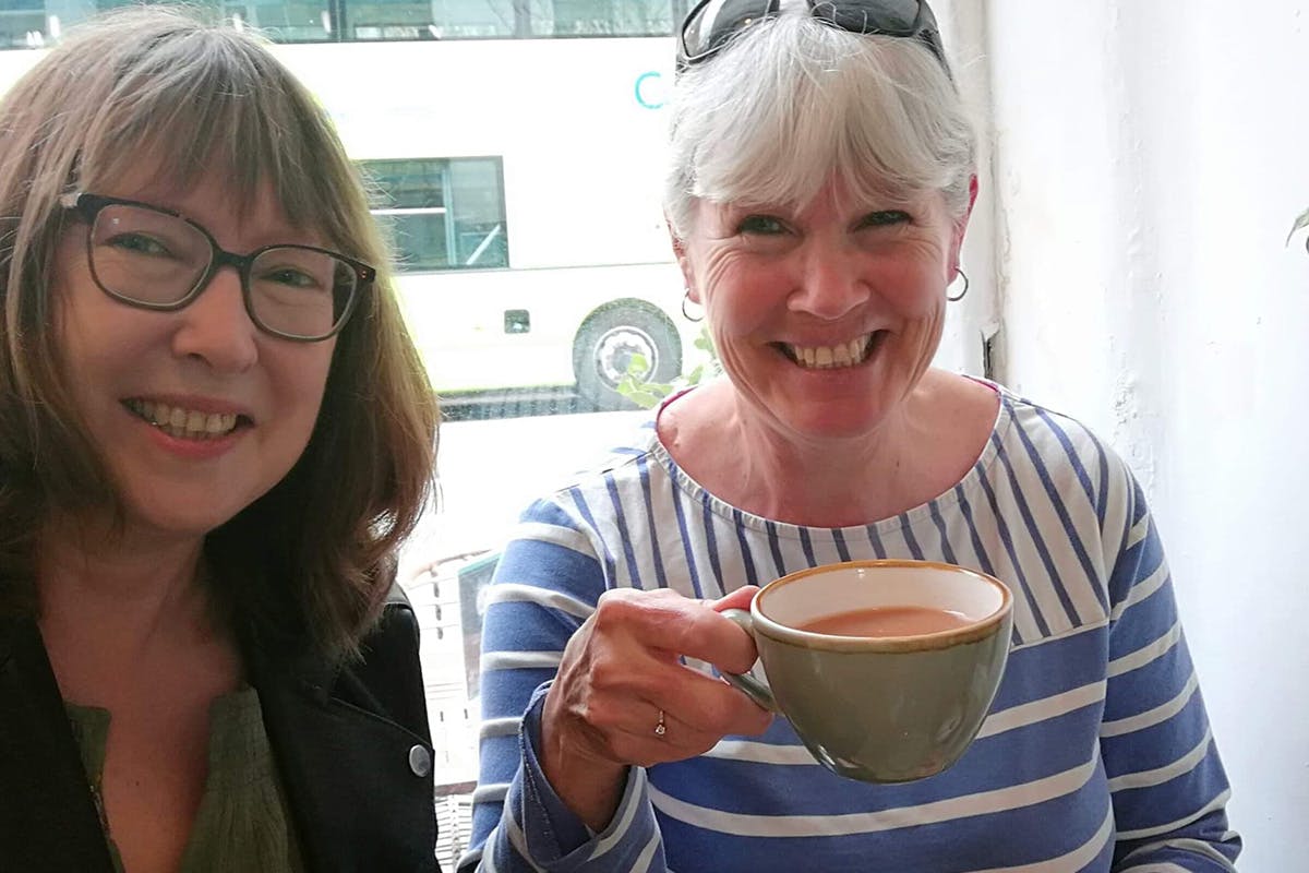 Two women smiling at the camera, one holding a full mug of tea