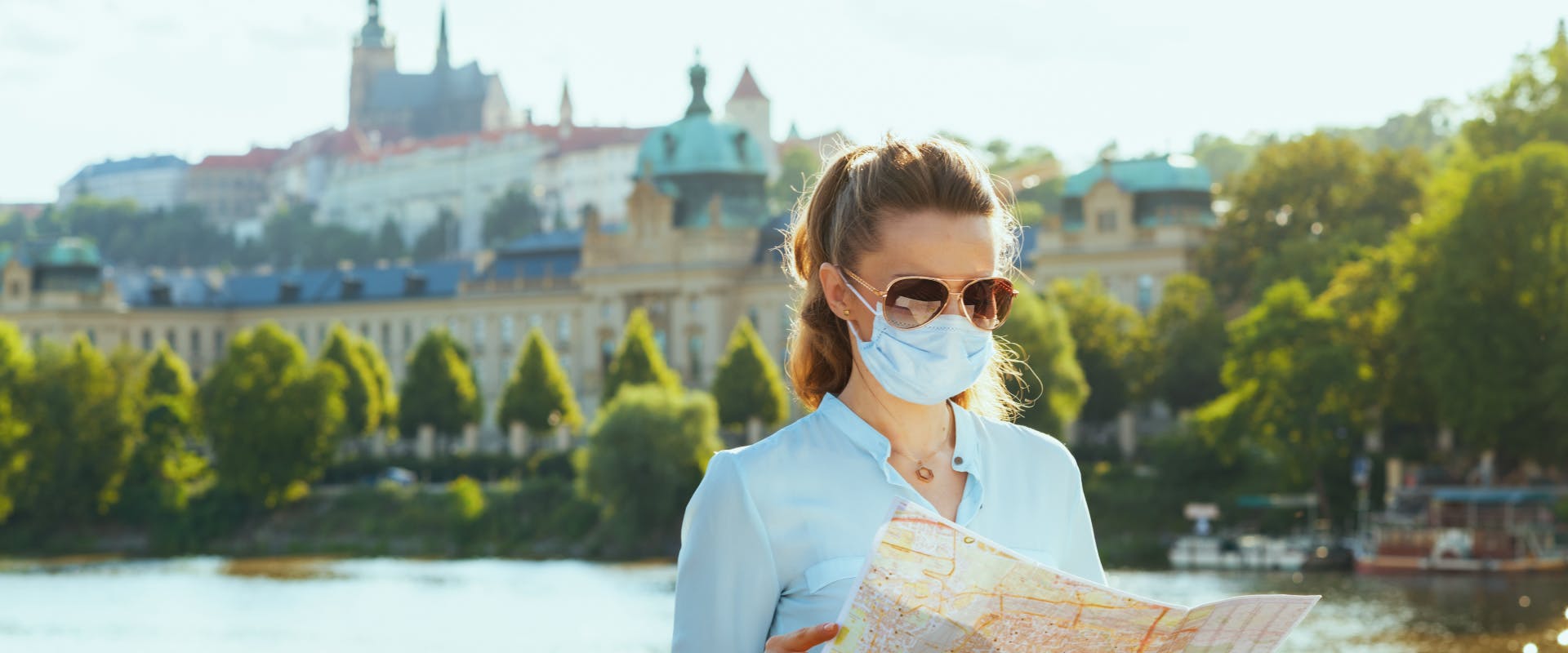 a solo female traveler wearing a mask whilst looking at a map with her back to Prague Castle and the Vltava River