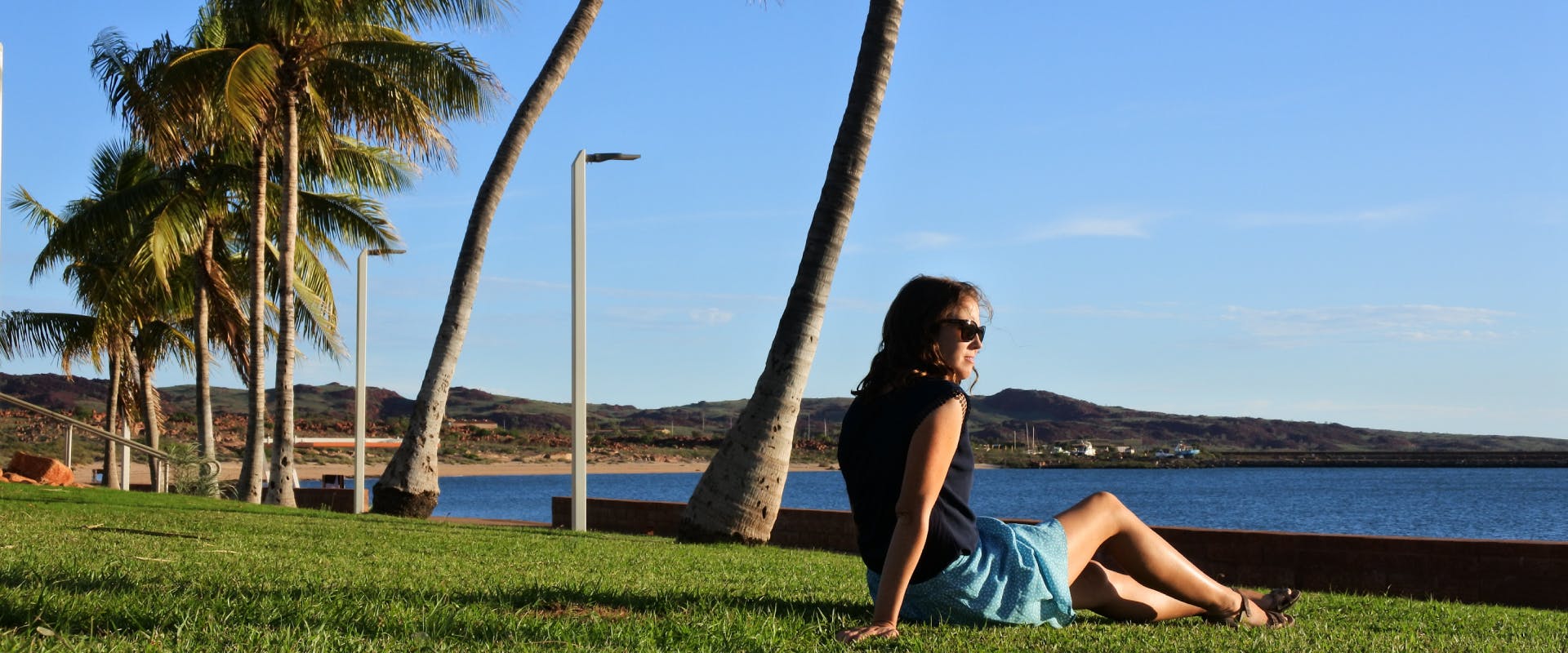 a coastline in australia with grass and palm trees and a solo female traveler sitting on the grass looking at the sea