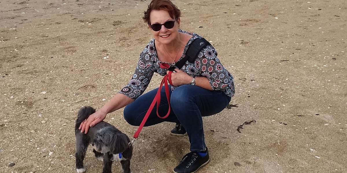 A woman on the beach smiling at the camera, petting a grey and white dog