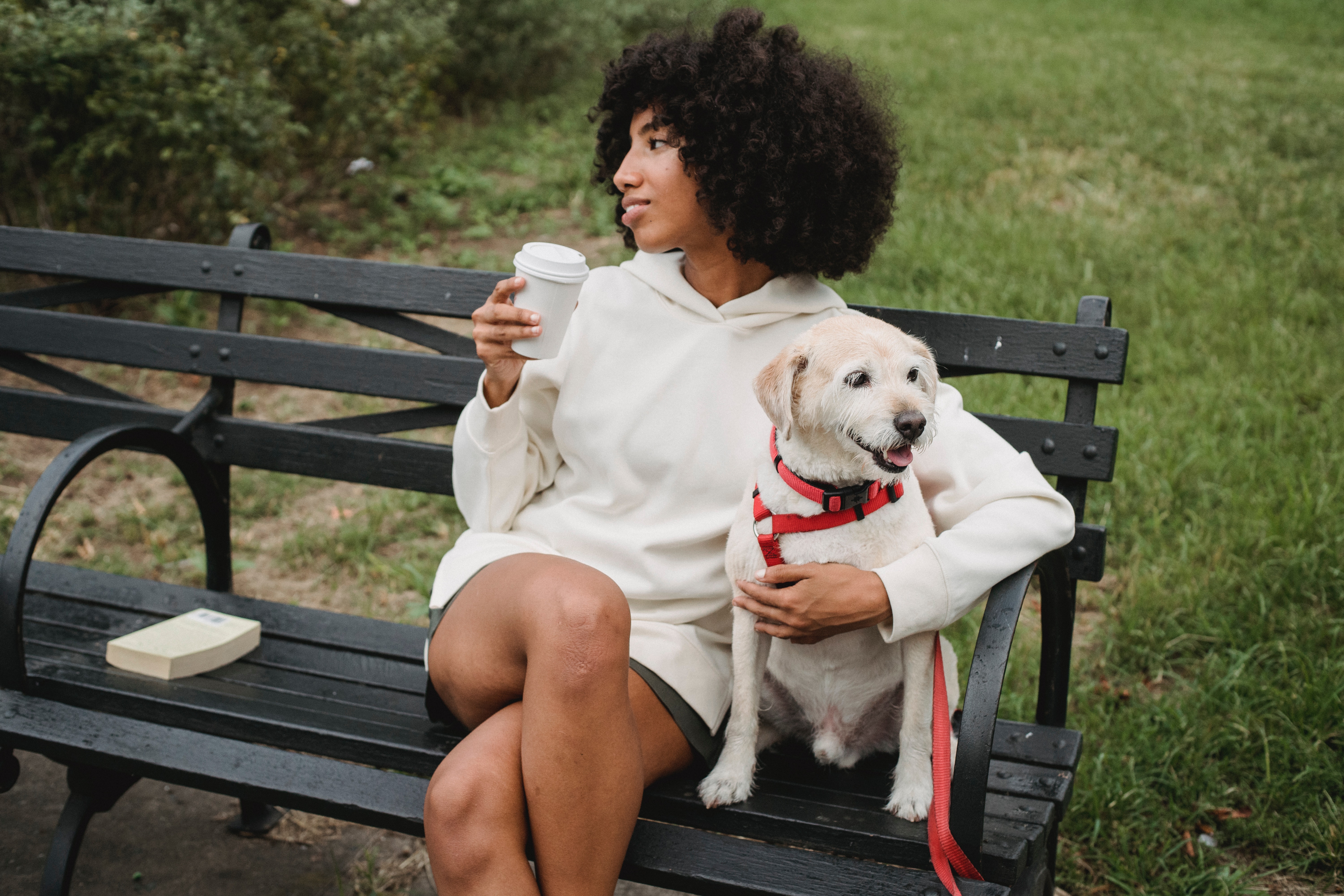 A woman sitting on a bench drinking a coffee, next to a small white dog