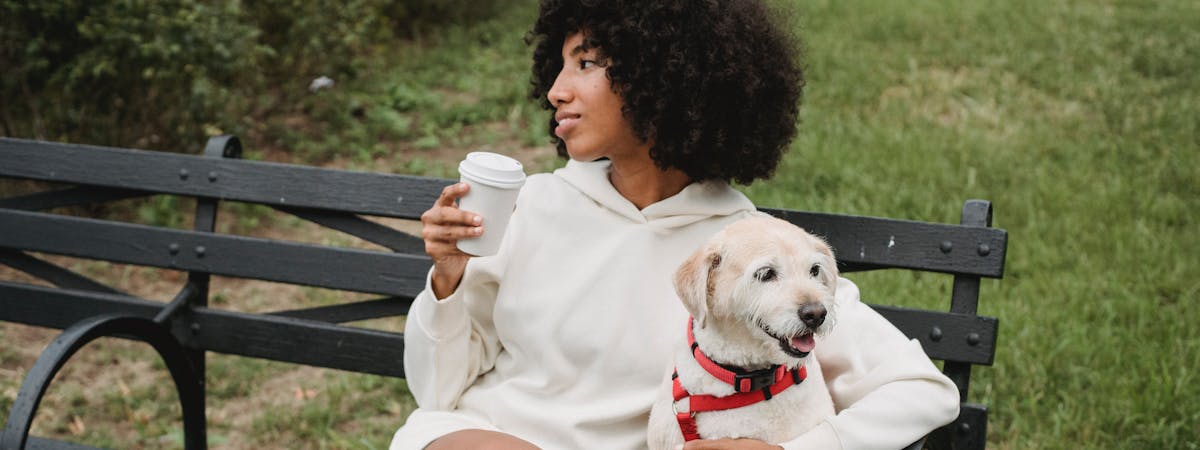 A woman sitting on a bench drinking a coffee, next to a small white dog