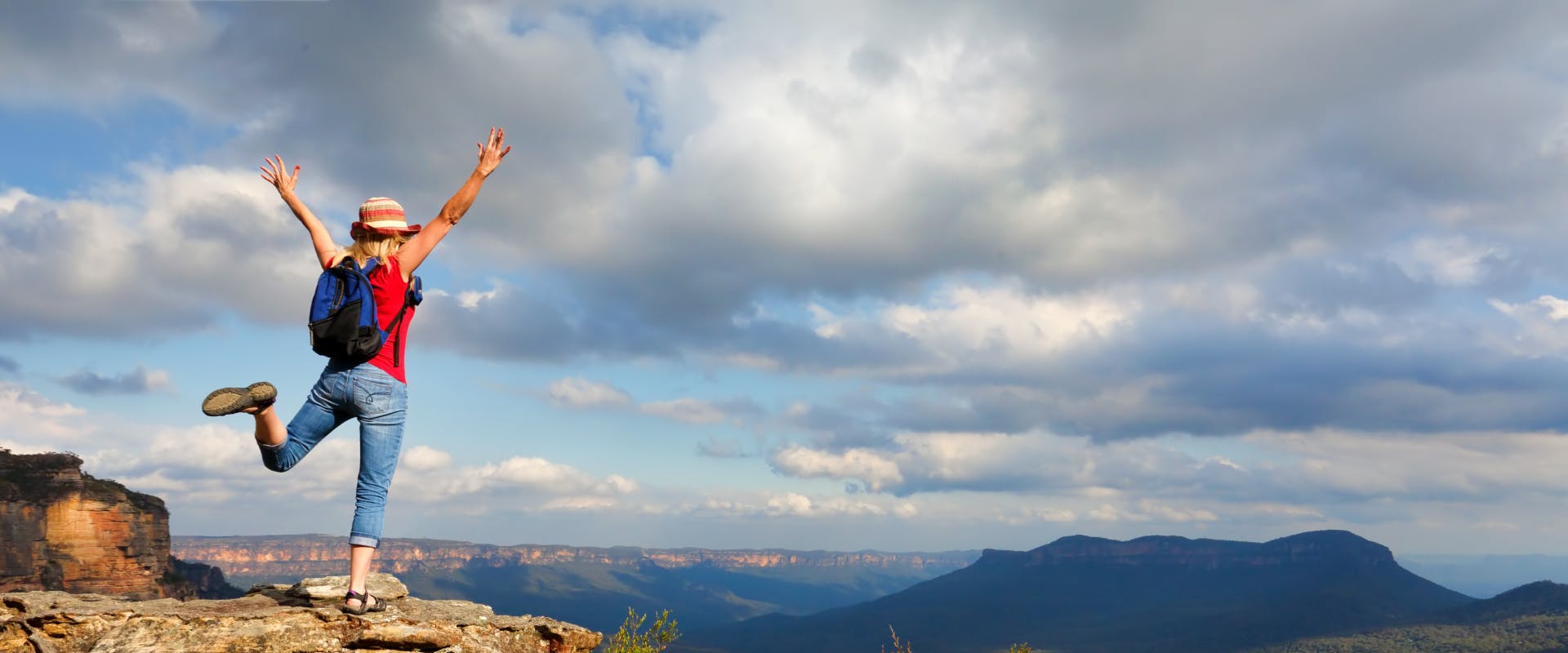 a solo female traveler jumping for joy on a cliff edge overlooking a rocky valley in australia