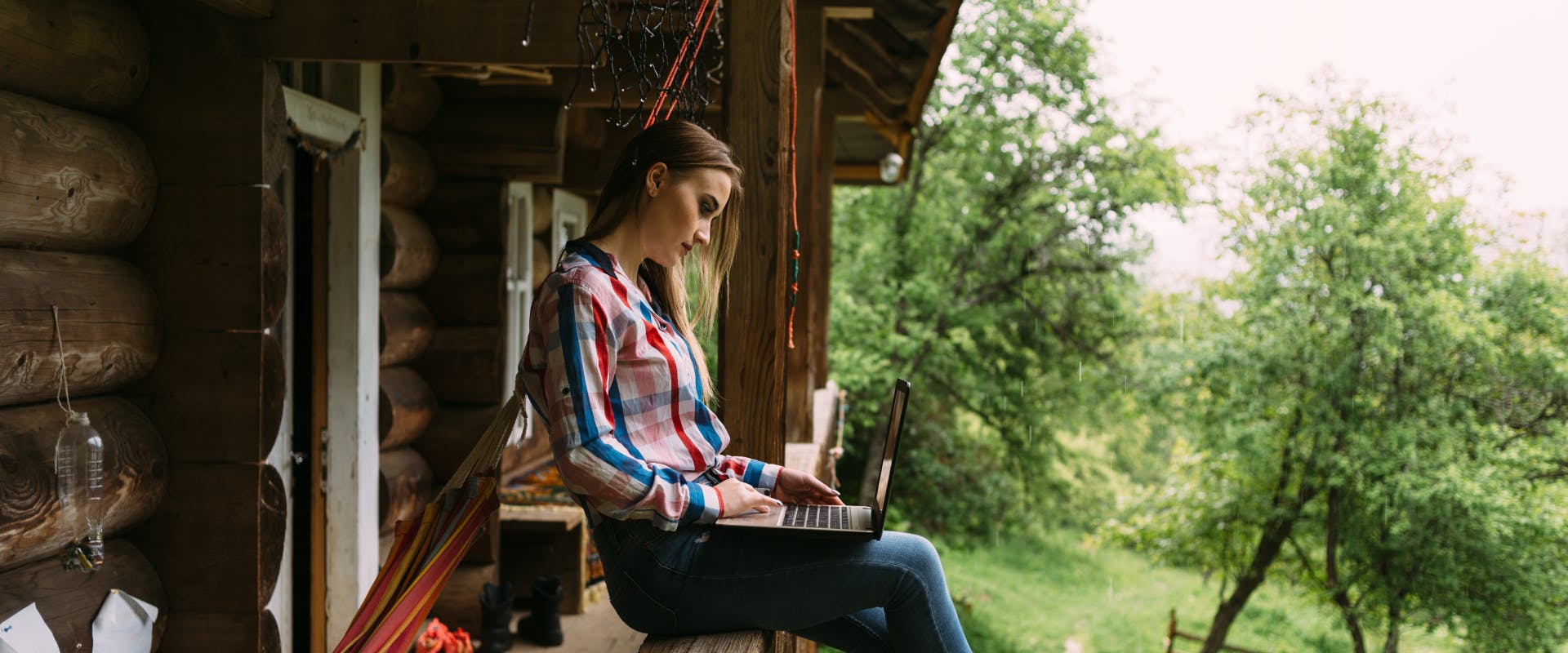 a woman sat on the porch fence of a log cabin whilst on her laptop in a rural area