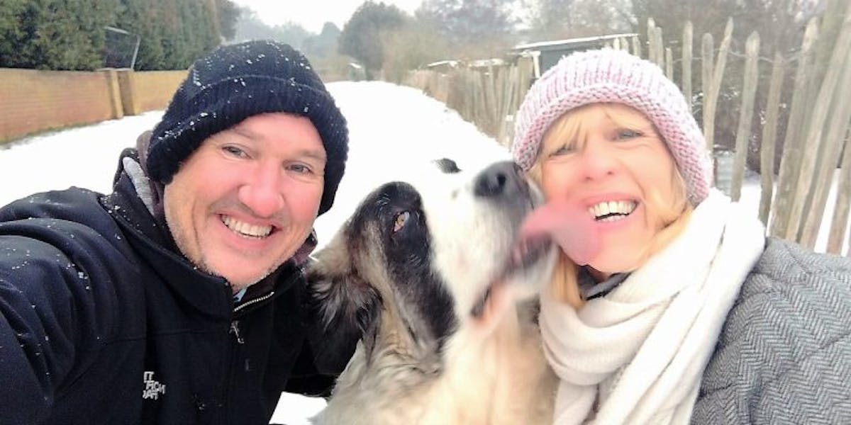 A man, a woman and a dog outside in the snow, the dog licking the woman's face