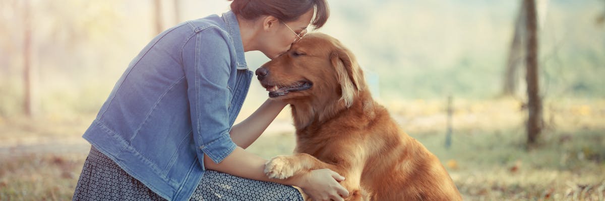 A woman in a forest kissing a dog on the top of its head 