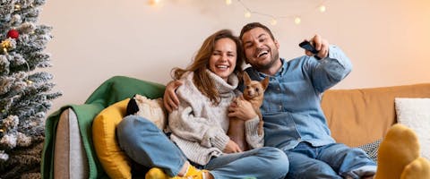 A couple and their dog sitting on the sofa, laughing at the television screen