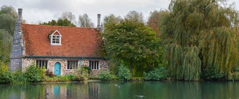 a stone cottage with a blue door on the banks of a large pond next to a willow tree in Colchester