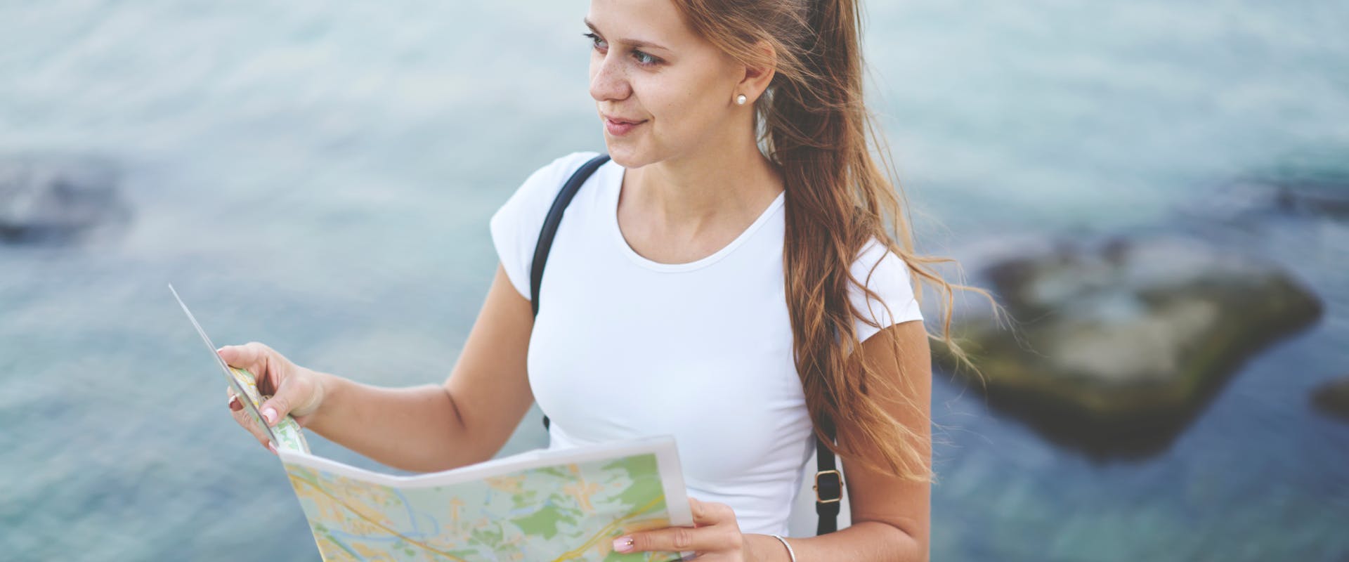 a solo female traveler next to a calm body of water looking up from a paper map