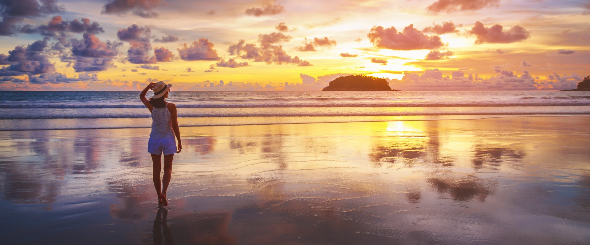 a solo female traveler walking along a beach at low tide with one hand on her sun hat watching the sun set