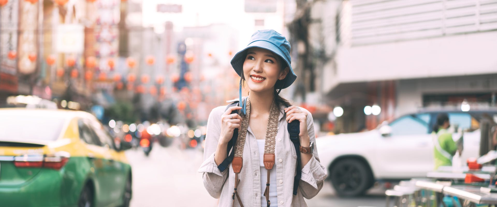 a solo female traveler walking down a busy street wearing a blue bucket hat and clutching the straps of her backpack while smiling
