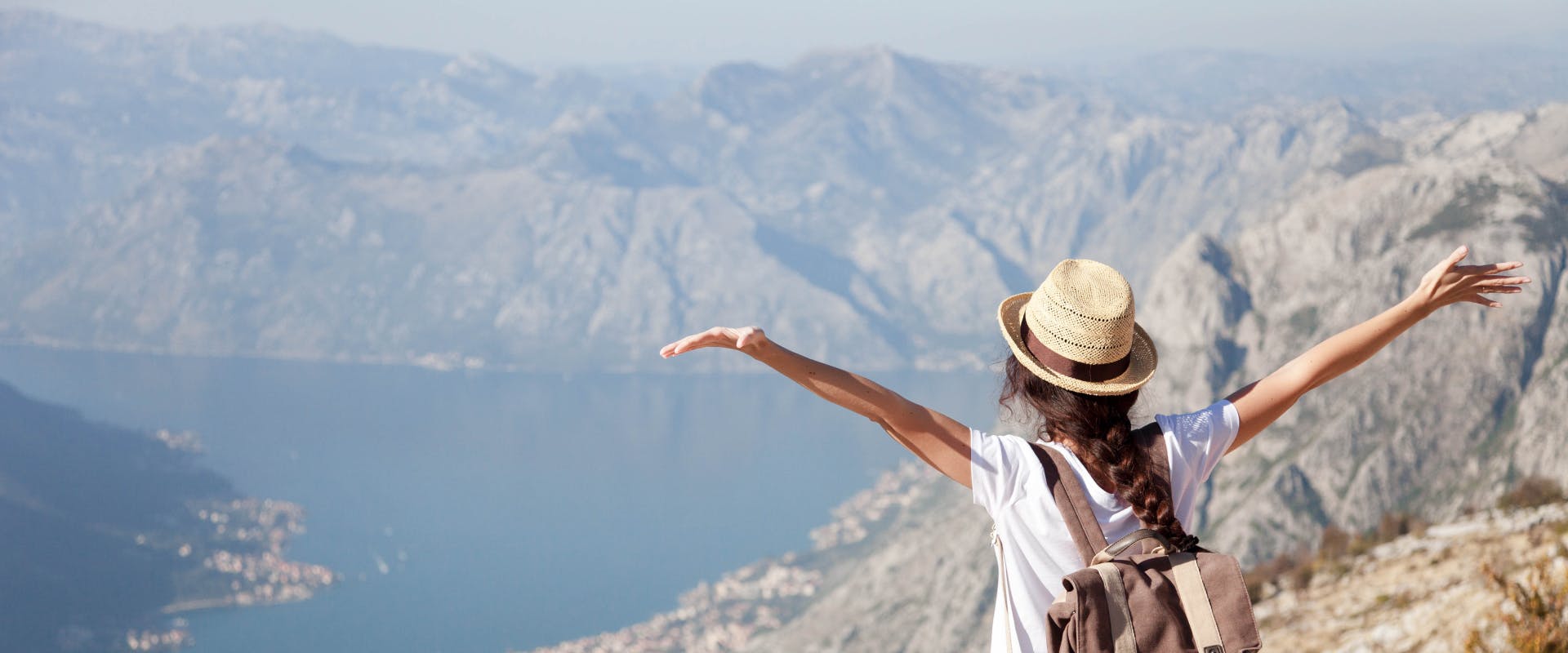 a solo female traveler looking over a valley lake at the top of a cliff with her arms lift upwards and outwards