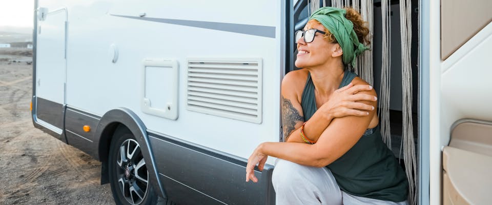 a solo female traveler wearing a green bandana and glasses sat in the doorway of a motorhome smiling at the sunset